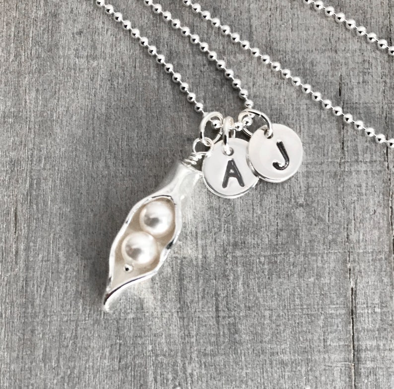 May include: Silver necklace with a pea pod charm and two pearl beads. The necklace has two initial charms, one with the letter 'A' and the other with the letter 'J'.