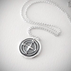 Puede incluir: Un collar de plata con un colgante de br&uacute;jula. La br&uacute;jula tiene un acabado plateado y presenta una rosa de los vientos con las letras N, S, E y W.
