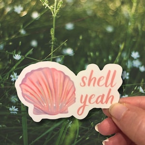 May include: A sticker featuring a pink seashell illustration with the words "shell yeah" in a matching pink script. The sticker is held against a blurred background of green foliage and white flowers.