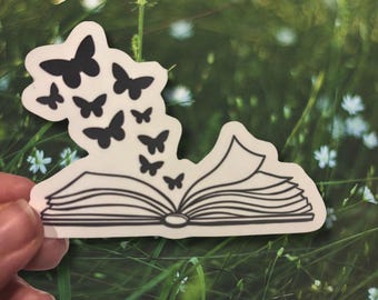 May include: A white sticker with a black outline of an open book with butterflies flying out of it. The sticker is on a green grassy background.