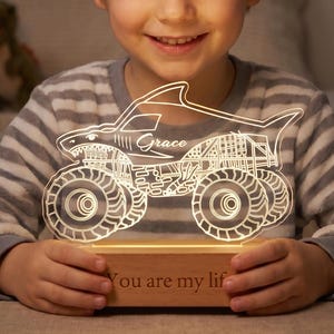 May include: A lit-up monster truck night light with the name "Grace" on the side. The truck is white and sits on a wooden base with the words "You are my lif" engraved. The boy is wearing a striped shirt.