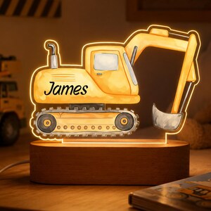 Könnte beinhalten: Ein gelbes LED-Nachtlicht in Form eines Baggers mit einem Holzsockel. Auf der Seite des Baggers steht der Name James in schwarzer Schreibschrift. Das Licht strahlt warm und betont das detaillierte Design des Baufahrzeugs und dessen Arm.