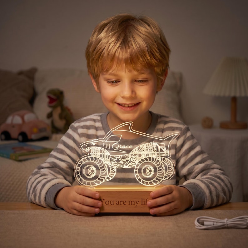 May include: A lit-up monster truck night light with the name "Grace" on the side. The truck is white and sits on a wooden base with the words "You are my lif" engraved. The boy is wearing a striped shirt.