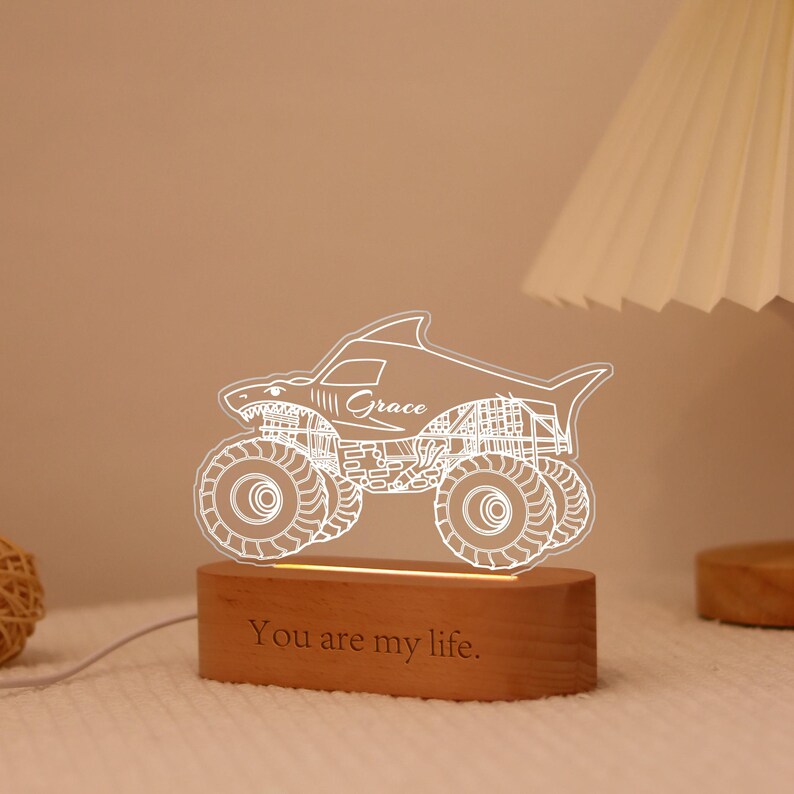 May include: A shark-shaped monster truck night light with the name "Grace" etched on the side. The light sits on a wooden base with the phrase "You are my life" engraved. The light is white and the base is brown.