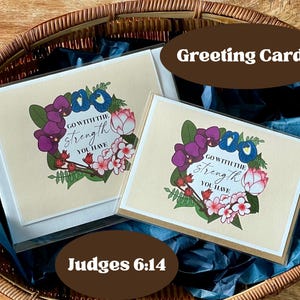 May include: Greeting cards with floral wreath designs. The cards feature a cream-colored background with a floral wreath in purple, blue, and pink. The text "GO WITH THE Strength YOU HAVE" is displayed. The cards are in a woven basket. The text "Judges 6:14" is also displayed.