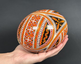 Puede incluir: Un huevo decorativo pintado a mano con intrincados patrones en naranja, rojo, negro y blanco. El huevo se sostiene en una mano, mostrando el detallado diseño de arte popular. La superficie del huevo es brillante, resaltando los colores vibrantes y los motivos geométricos.