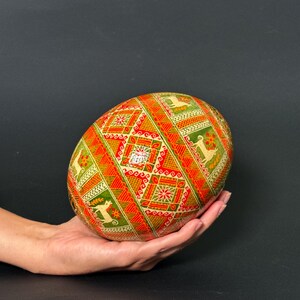 May include: A decorative, patterned egg held in a hand. The egg features a geometric design with red, green, and gold colors. The pattern includes stylized animal figures and floral motifs, creating a festive and detailed appearance.