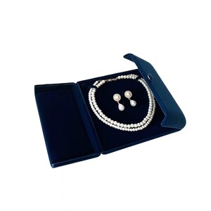 Op de afbeelding: Een parelketting met twee strengen en bijpassende pareloorbellen, gepresenteerd in een donkerblauwe fluwelen sieradendoos. De oorbellen hebben een pareldruppelontwerp. De sieradendoos is open en onthult de witte parels tegen de donkerblauwe binnenkant.