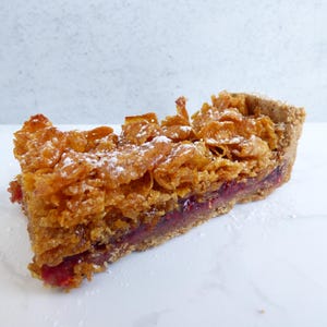 May include: A slice of dessert with a crispy, golden brown cereal crust and a layer of red jam. The crust is dusted with icing sugar.