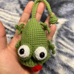 May include: A handmade green crocheted creature with large white-rimmed eyes, black pupils, and a red mouth. It has small arms and a curved stem with a ball at the end. The whimsical design is likely a decorative item or toy.