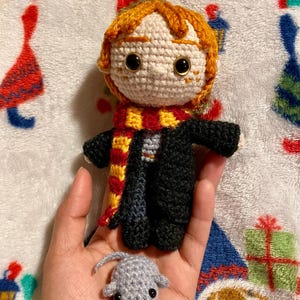 May include: A crocheted Harry Potter doll with a red and gold scarf, black robe, and brown hair. The doll is holding a small gray crocheted rat.