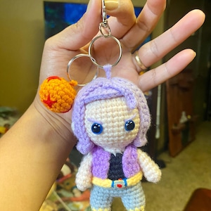 May include: Crocheted keychain featuring a character with purple hair, a purple vest, and blue pants. The keychain also includes a small orange ball with a red star. The character has blue eyes and is attached to a silver keyring.