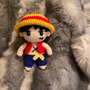 May include: Crocheted figure of a cartoon character wearing a red and yellow hat, red vest, and blue pants. The figure is sitting on a gray fuzzy surface.