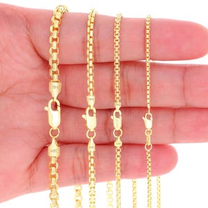 May include: Five gold chains of varying lengths, displayed on a hand. The chains are all box chains, with a smooth, polished finish.