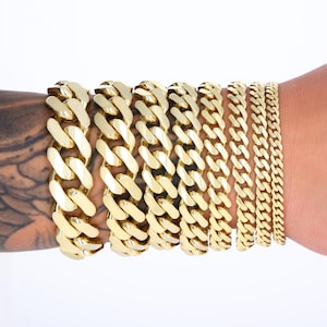 May include: A set of gold-tone chain bracelets of varying widths, stacked on a person's wrist.
