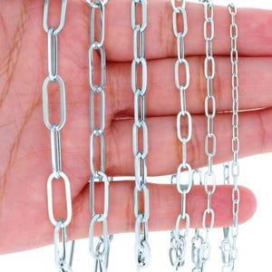 May include: Six silver-colored chain necklaces with varying link sizes are displayed. The chains are held in a hand, showcasing the different styles and lengths. Each chain has a clasp for secure wear. The links are oval and rectangular.