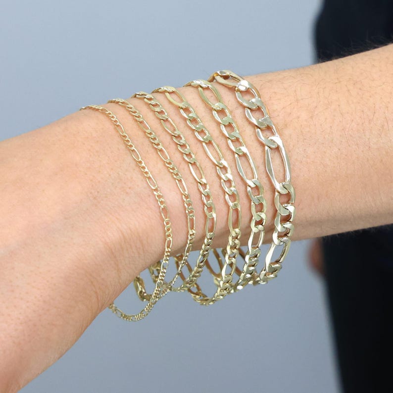 May include: A set of six gold chain bracelets of varying widths, stacked on a person's wrist.