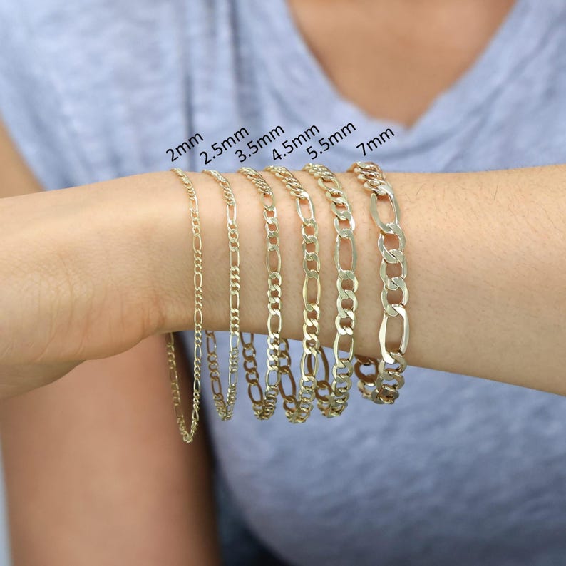 May include: A close-up of a person's wrist wearing six gold chain bracelets of different widths. The bracelets are labelled with their widths in millimetres: 2mm, 2.5mm, 3.5mm, 4.5mm, 5.5mm, and 7mm.