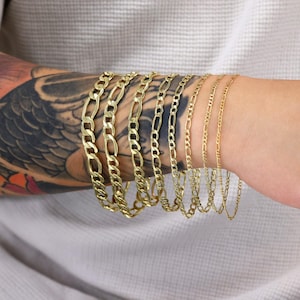 May include: A set of gold chain bracelets of varying widths, stacked on a person's arm. The chains are all made of a classic, flat, curb link style.