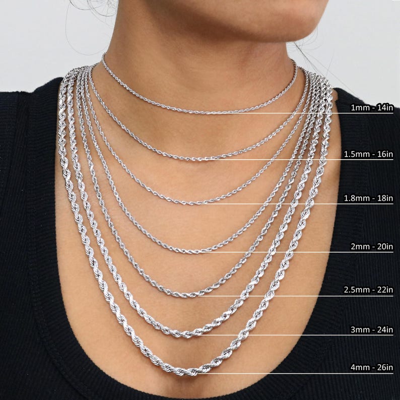 May include: A close-up of a model wearing a variety of silver rope chains of different lengths and thicknesses. The chains are labelled with their thickness and length in inches. 1mm - 14in, 1.5mm - 16in, 1.8mm - 18in, 2mm - 20in, 2.5mm - 22in, 3mm - 24in, 4mm - 26in.