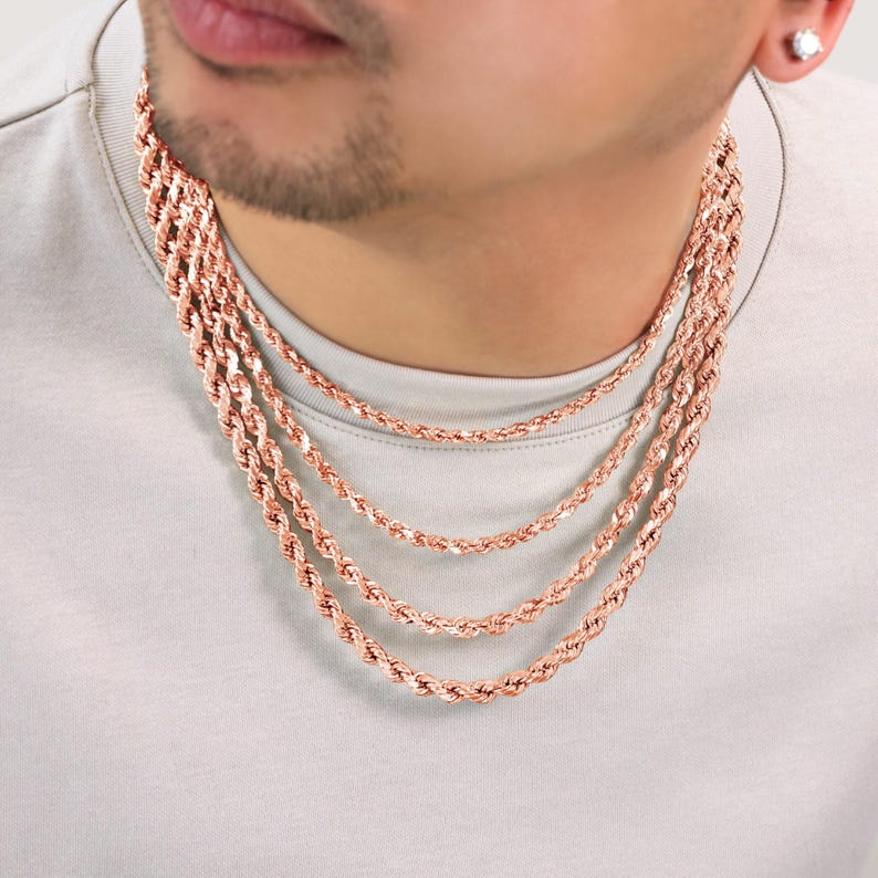 Puede incluir: Un collar de cadena de cuerda de oro rosa en capas. La cadena est&aacute; hecha de m&uacute;ltiples hebras de cadena de cuerda, cada una con un grosor diferente. El collar lo lleva una persona con una camisa marr&oacute;n claro.