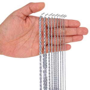 May include: A close-up of a hand holding a variety of silver rope chains. The chains are arranged in order of size, with the largest chain at the top and the smallest chain at the bottom. The chains are labelled with their respective sizes in millimetres: 5mm, 4mm, 3.5mm, 3mm, 2.5mm, 2mm, 1.8mm, 1.5mm, and 1mm.