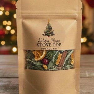 May include: A kraft paper pouch with a clear window, labeled "Holiday Magic Stove Top Potpourri." The pouch contains dried orange slices, cinnamon sticks, and greenery. A Christmas tree graphic is on the label. The background is blurred with Christmas lights.
