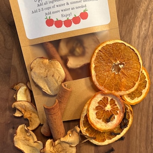 May include: A brown paper bag with a white label that reads "Apple Pie Simmer Pot" and instructions for making a simmer pot. The bag contains dried apple slices, cinnamon sticks, orange slices, and cloves.