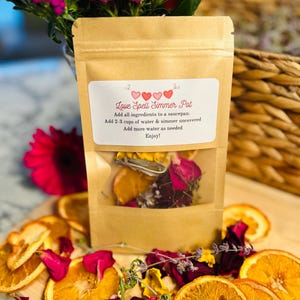 May include: A brown paper bag with a clear window showing dried orange slices, rose petals, and lavender. The bag has a label that reads "Love Spell Simmer Pot" and instructions for use.