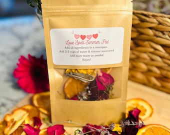 Op de afbeelding: Een bruine papieren zak met een doorzichtig venster dat gedroogde sinaasappelschijfjes, rozenblaadjes en lavendel laat zien. De zak heeft een label met de tekst "Love Spell Simmer Pot" en gebruiksaanwijzingen.