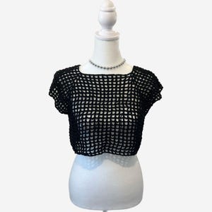 May include: Black crochet crop top with a square neckline and short sleeves. The top is displayed on a white mannequin. A silver beaded necklace is around the mannequin's neck. The top is made of a net-like material.