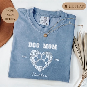 Puede incluir: Una camiseta azul vaquera Comfort Colors con el texto "DOG MOM" y un diseño de corazón con huella de pata. La camiseta también incluye el nombre "Charlie" y el año "2020". Un collar dorado está drapeado sobre la camiseta.