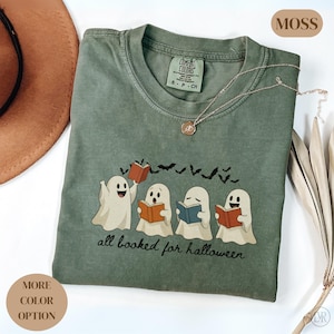 Puede incluir: Una camiseta de manga corta color verde musgo con un gráfico de cuatro fantasmas blancos leyendo libros coloridos. Sobre los fantasmas hay siluetas negras de murciélagos volando. El texto "all booked for halloween" está impreso en negro debajo de los fantasmas.