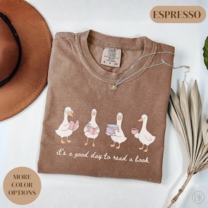 Puede incluir: Camiseta Comfort Colors color Espresso con un gráfico de cuatro gansos de dibujos animados, cada uno con un libro o una taza de café. El texto "it's a good day to read a book" está impreso debajo. Un collar plateado está sobre la camiseta.