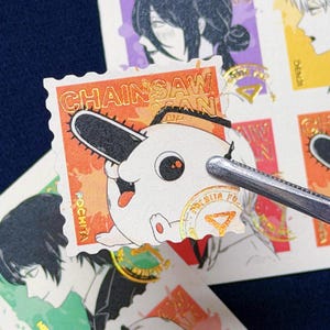 Puede incluir: Una hoja de pegatinas con personajes de anime. Una pegatina muestra una criatura blanca con una cabeza de motosierra, el texto "CHAINSAW MAN" y "POCHITA". Otras pegatinas presentan personajes de anime en varios colores.