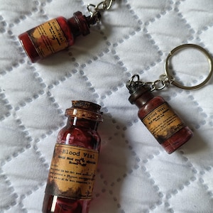 Könnte beinhalten: Drei Miniatur-Glasfläschchen, gefüllt mit einer roten Flüssigkeit, mit der Aufschrift "Blood Vial", sind an Schlüsselanhängern befestigt. Die Fläschchen haben Korken und alt aussehende Etiketten. Der Hintergrund ist ein weißer, gesteppter Stoff.
