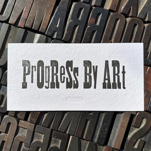 Op de afbeelding: Een witte kaart met de tekst "Progress By Art" gedrukt in zwarte letters op een achtergrond van vintage houten letterpress blokken.