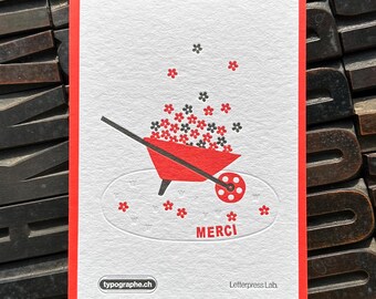 May include: A red and white greeting card with a red border. The card features a red wheelbarrow spilling red and black flowers. The word "MERCI" is printed below the wheelbarrow. The website "typographe.ch" is printed on the bottom left.