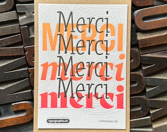 May include: A card with the word "Merci" printed in orange, red, and black on a white background. The card is surrounded by wooden letterpress blocks.