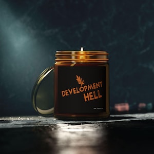 May include: A brown glass jar candle with a gold lid and a black label. The label features the words "DEVELOPMENT HELL" in orange, along with a small leaf design. The candle is lit, and the lid is partially open.
