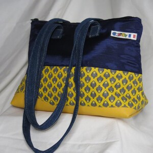 Peut inclure: Un sac fourre-tout avec un dessus bleu marine et des poignées en jean. Le bas est jaune avec un motif jaune et gris. Le sac a une petite étiquette avec du texte et un drapeau.