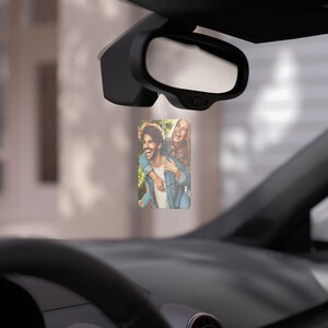 Può includere: Profumatore per auto rettangolare con una foto di una coppia sorridente, appeso allo specchietto retrovisore di un'auto. La foto mostra un uomo che porta una donna sulla schiena, entrambi con giacche di jeans.