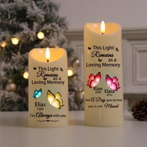 Puede incluir: Dos velas LED de color marfil con una llama parpadeante. Cada vela tiene un diseño de mariposa y el texto "This Light Remains as a Loving Memory". Una vela dice "Elias 1969 I'm Always with you". La otra dice "Kian 2008 Elara 2012 Not A Day goes by that you're not Missed."