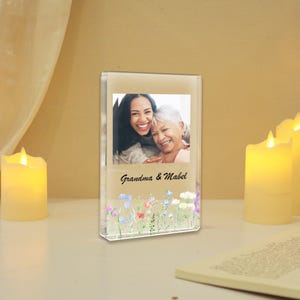 Puede incluir: Un bloque de fotos de acrílico transparente con una foto de dos mujeres sonriendo, con el texto "Grandma & Mabel" debajo. La parte inferior del bloque tiene un diseño floral con flores coloridas. El bloque está sobre una superficie blanca con velas encendidas.
