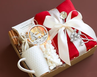May include: A gift basket filled with a white mug, a bag of marshmallows, a wooden reindeer ornament, cinnamon sticks, and a box of caramels. The basket is wrapped in red fabric with a white ribbon and silver decorative sprigs.