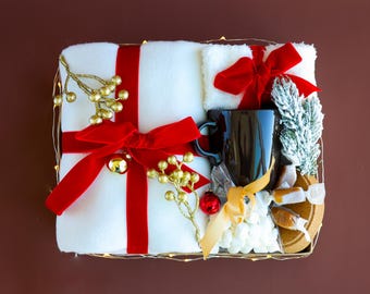 May include: A white blanket, red ribbon, and gold ornaments are arranged in a basket with a black mug, marshmallows, and caramel candies. This gift basket is perfect for a cozy night in.