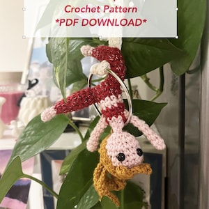 May include: A crochet figure of an acrobat hanging from a silver metal hoop. The figure has a light pink head, a red and white striped body, and a mustard yellow lower section. Text overlay reads: Mini Aerial Hoop Acrobat Crochet Pattern *PDF DOWNLOAD*