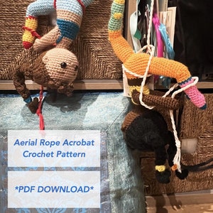 Può includere: Due acrobati aerei in crochet appesi a uno scaffale. L'acrobata a sinistra indossa un body blu e viola con una striscia gialla e rossa. L'acrobata a destra indossa un body arancione e giallo con una striscia rosa. Il testo "Aerial Rope Acrobat Crochet Pattern" e "*PDF DOWNLOAD*" è visibile sotto gli acrobati.