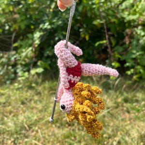 May include: A crocheted figure with light pink skin and curly golden-yellow hair, wearing a red and white patterned leotard. The figure is attached to a silver-colored metal pole, positioned in a pose that mimics a pole dance performance.