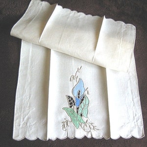 May include: Cream-colored linen towel with a scalloped edge, featuring an embroidered blue and green floral design. A folded section of the towel is visible on top. The towel is likely for the bathroom or kitchen.